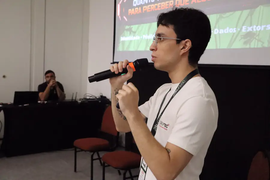 Palestrante de camiseta branca fala ao microfone diante do telão.